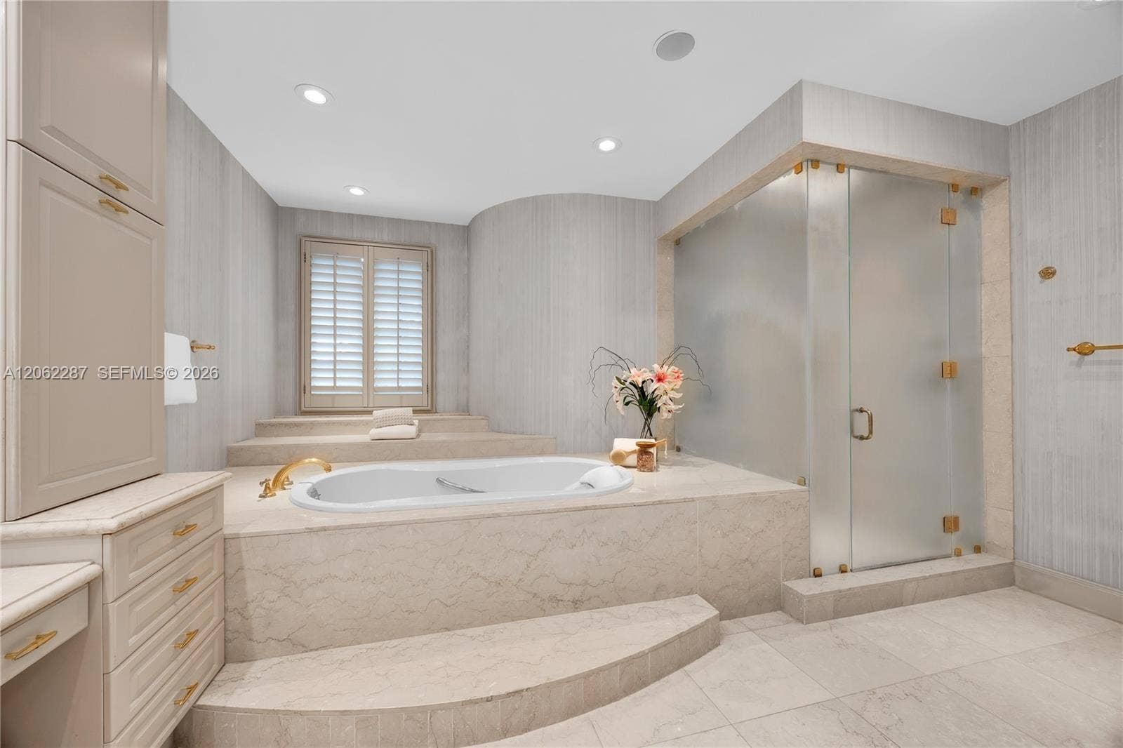 Raised stone-surround soaking tub with gold-tone fixtures, walk-in glass-enclosed shower, custom built-in cabinetry, floor-to-ceiling stone wall cladding, and recessed lighting