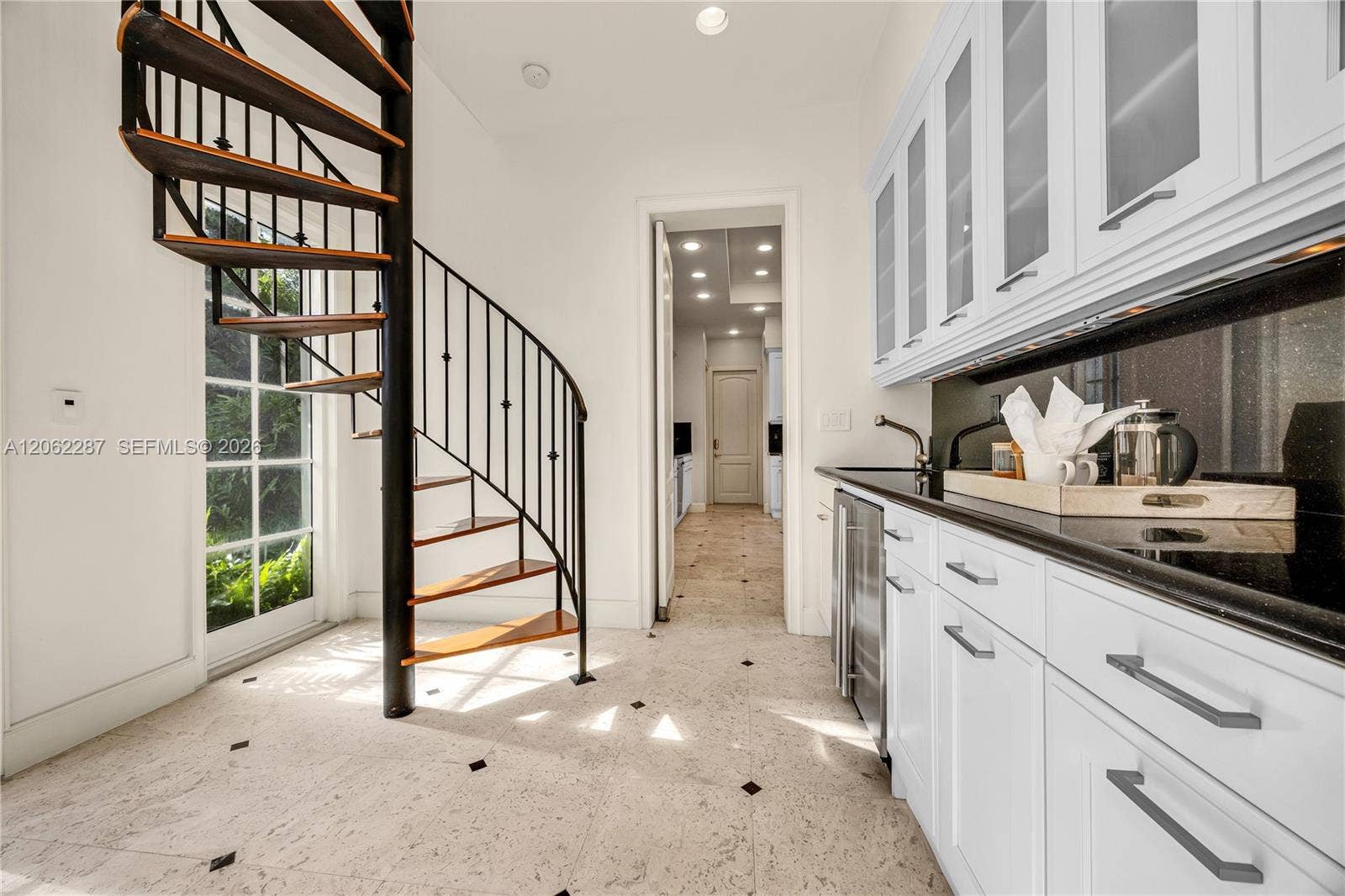 Spiral staircase with metal railing and wood-finish treads, custom white cabinetry featuring glass-front upper doors, dark stone-finish countertops, stainless steel under-counter refrigerator, and light-toned tile flooring with dark insets