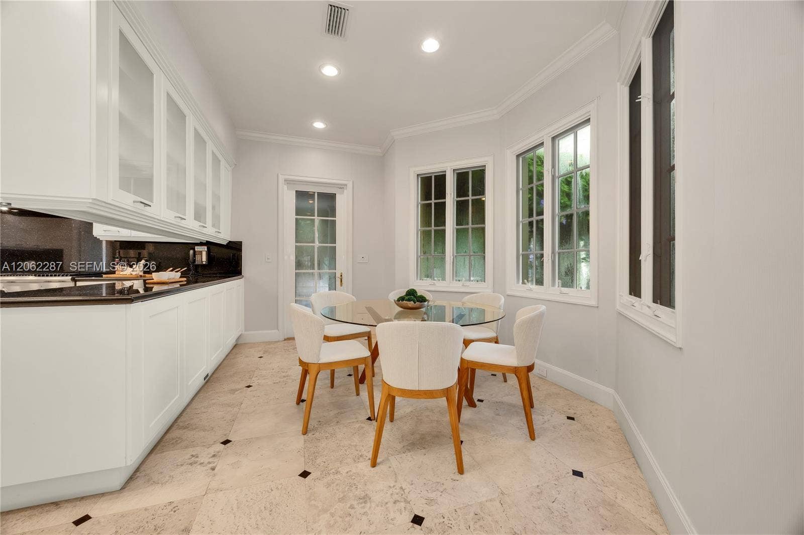 Custom white cabinetry with glass-panel upper doors, dark stone countertops, recessed lighting, multi-pane windows, and tile flooring with decorative stone insets