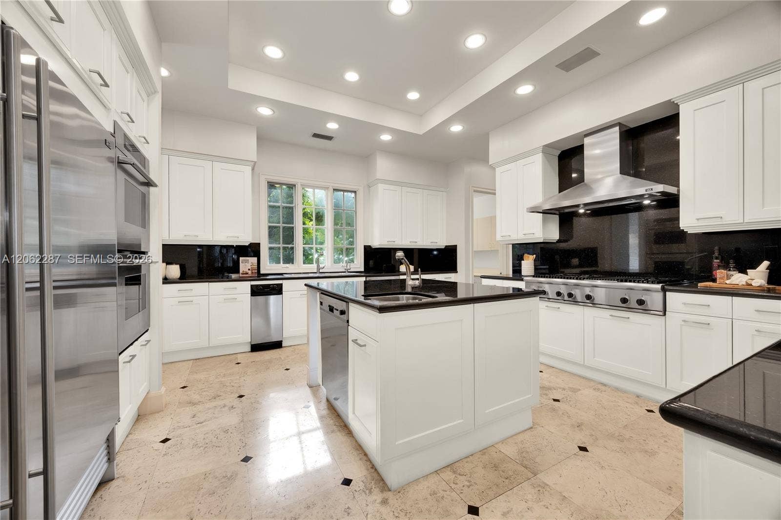 Kitchen featuring white shaker-style cabinetry, a central island with a stone countertop, stainless steel appliances, a professional-grade range hood, and stone flooring with inset decorative tile accents