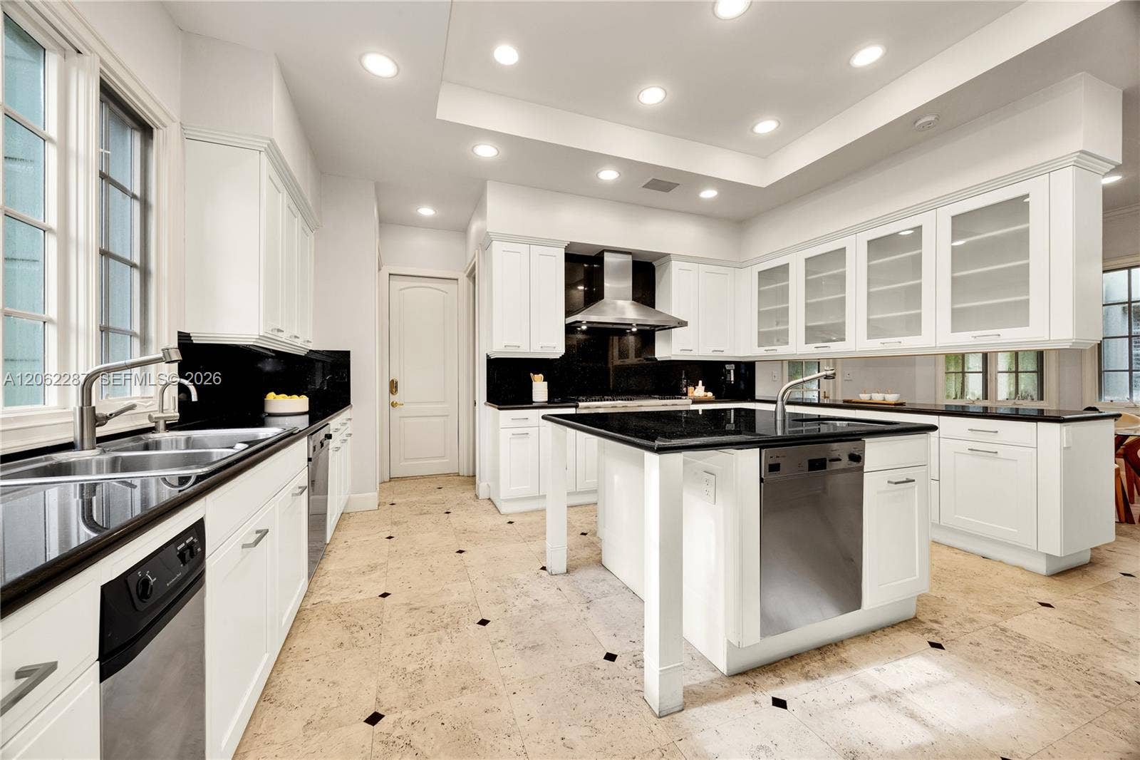 Kitchen featuring white shaker-style cabinetry, contrasting black countertops, a central island with integrated stainless steel dishwasher, recessed ceiling lighting, and stone flooring with inlaid accents