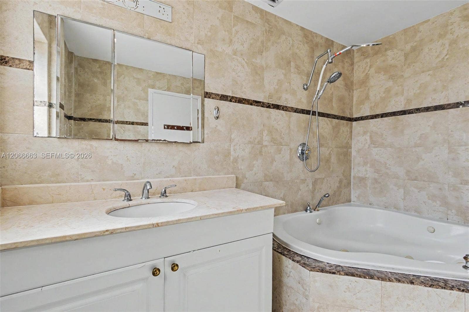 1191 NE 175th Street, North Miami Beach, FL 33162 Bathroom featuring a white vanity cabinet, light stone countertop with an integrated basin, triple-panel mirrored medicine cabinet, built-in jetted tub, and wall tiling with decorative border accent