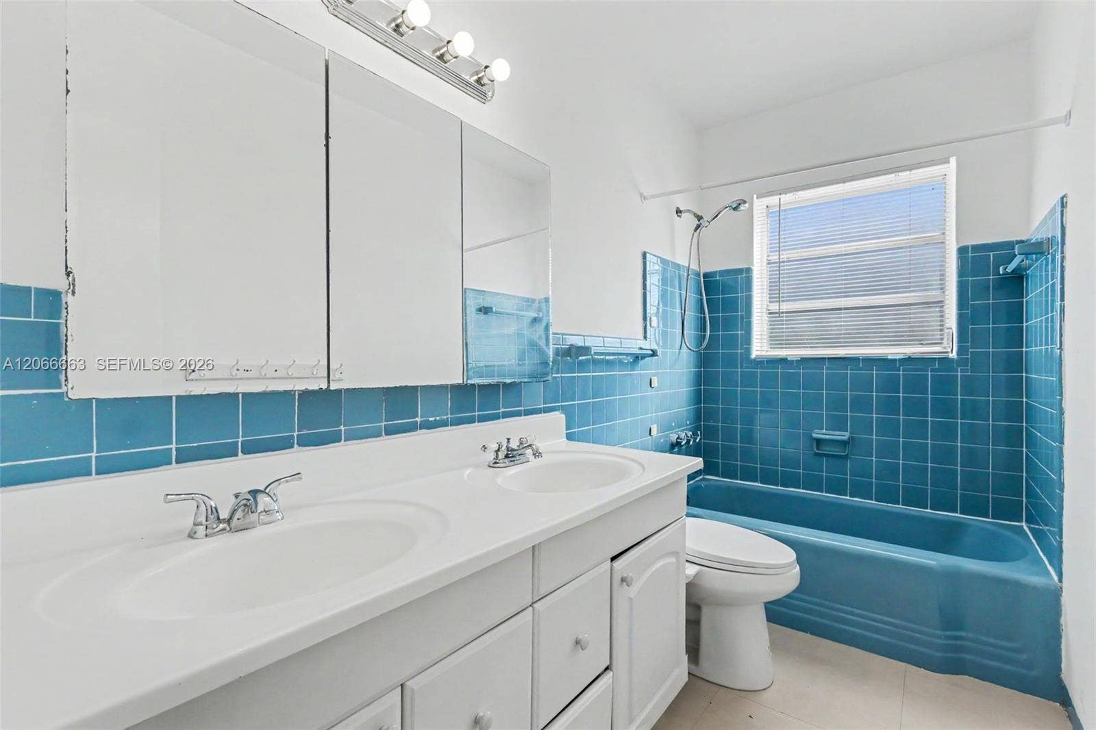 1191 NE 175th Street, North Miami Beach, FL 33162 Dual-sink vanity with white countertop, integrated medicine cabinet mirrors, multi-bulb wall lighting, and blue tile wainscoting surrounding the matching blue bathtub