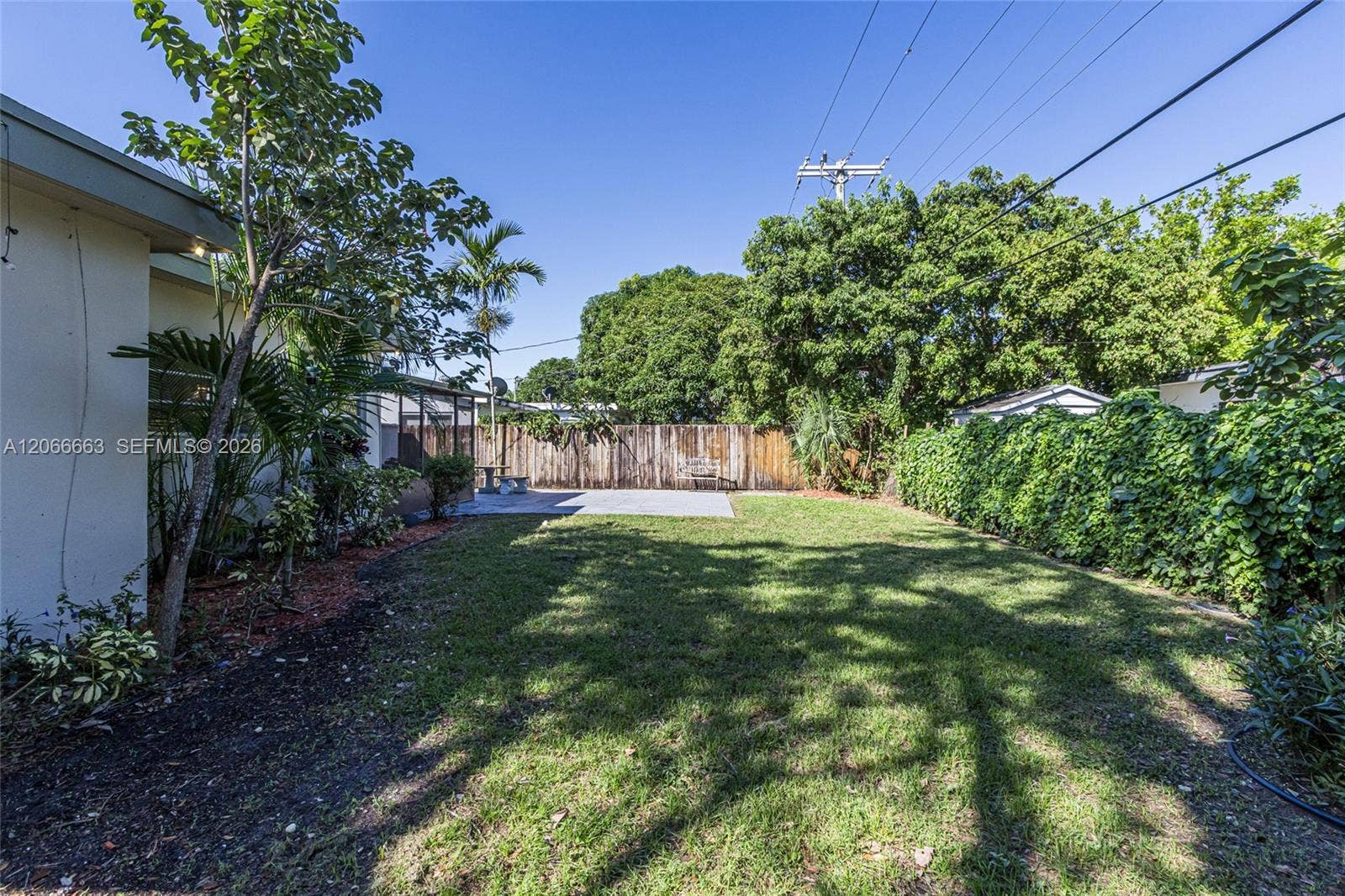 1191 NE 175th Street, North Miami Beach, FL 33162 Expansive backyard featuring a manicured lawn, concrete patio, mature trees, and wooden perimeter fencing