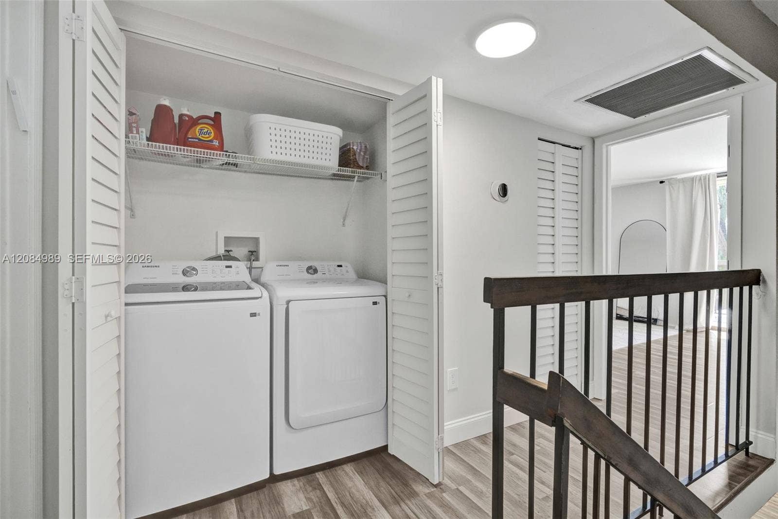 Laundry area featuring louvered bi-fold closet doors, integrated wire shelving, and wood-finish flooring with a metal stair railing