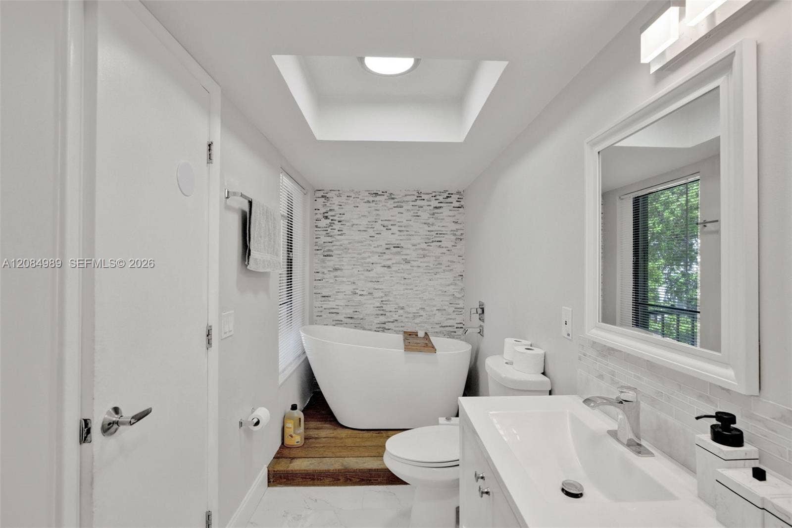 Freestanding soaking tub set upon a wood-finish platform, stone-tile accent wall, recessed ceiling lighting, and a white vanity with integrated sink