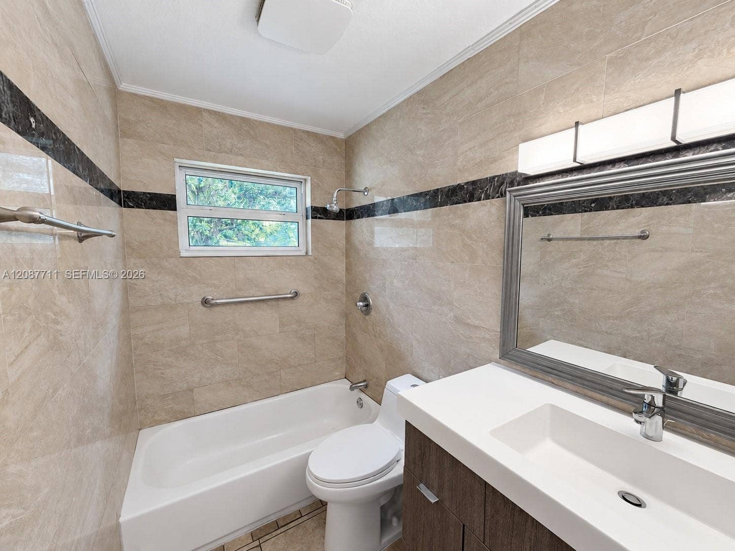 80 NE 162nd Street, Miami, Miami, FL 33162 Full-height neutral stone tiling with decorative dark stone accent banding, modern wood-finish vanity with integrated rectangular sink, wall-mounted multi-light fixture, and built-in grab bars