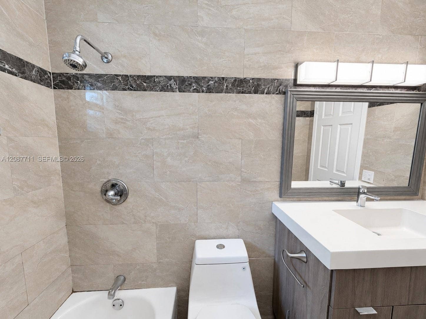 80 NE 162nd Street, Miami, Miami, FL 33162 Bathroom featuring floor-to-ceiling tile with a contrasting decorative inlay, a wood-finish vanity with a white countertop and integrated sink, a contemporary multi-light fixture, and a framed mirror