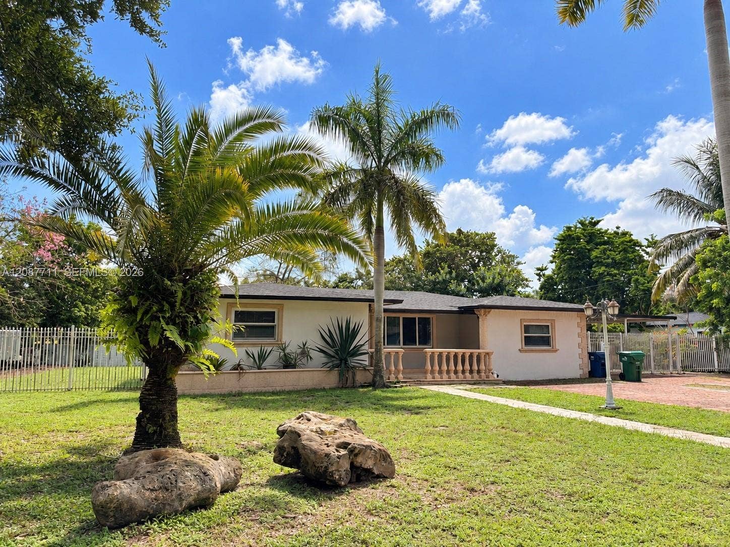 80 NE 162nd Street, Miami, Miami, FL 33162 Single-story residence featuring a light-colored stucco exterior, decorative stone balustrade, mature palm trees, manicured lawn, and perimeter metal fencing