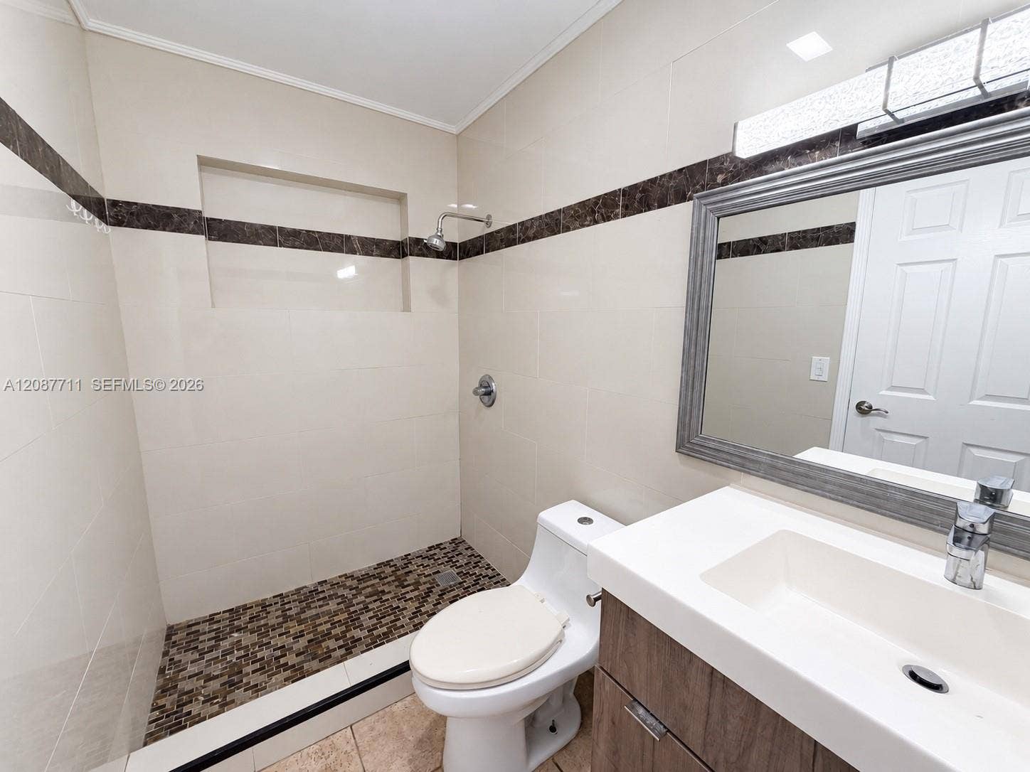 80 NE 162nd Street, Miami, Miami, FL 33162 Bathroom featuring large-format wall tile with decorative stone inlay, a walk-in shower with stone mosaic flooring and built-in recessed shelving, and a vanity with a white integrated sink and wood-finish cabinetry