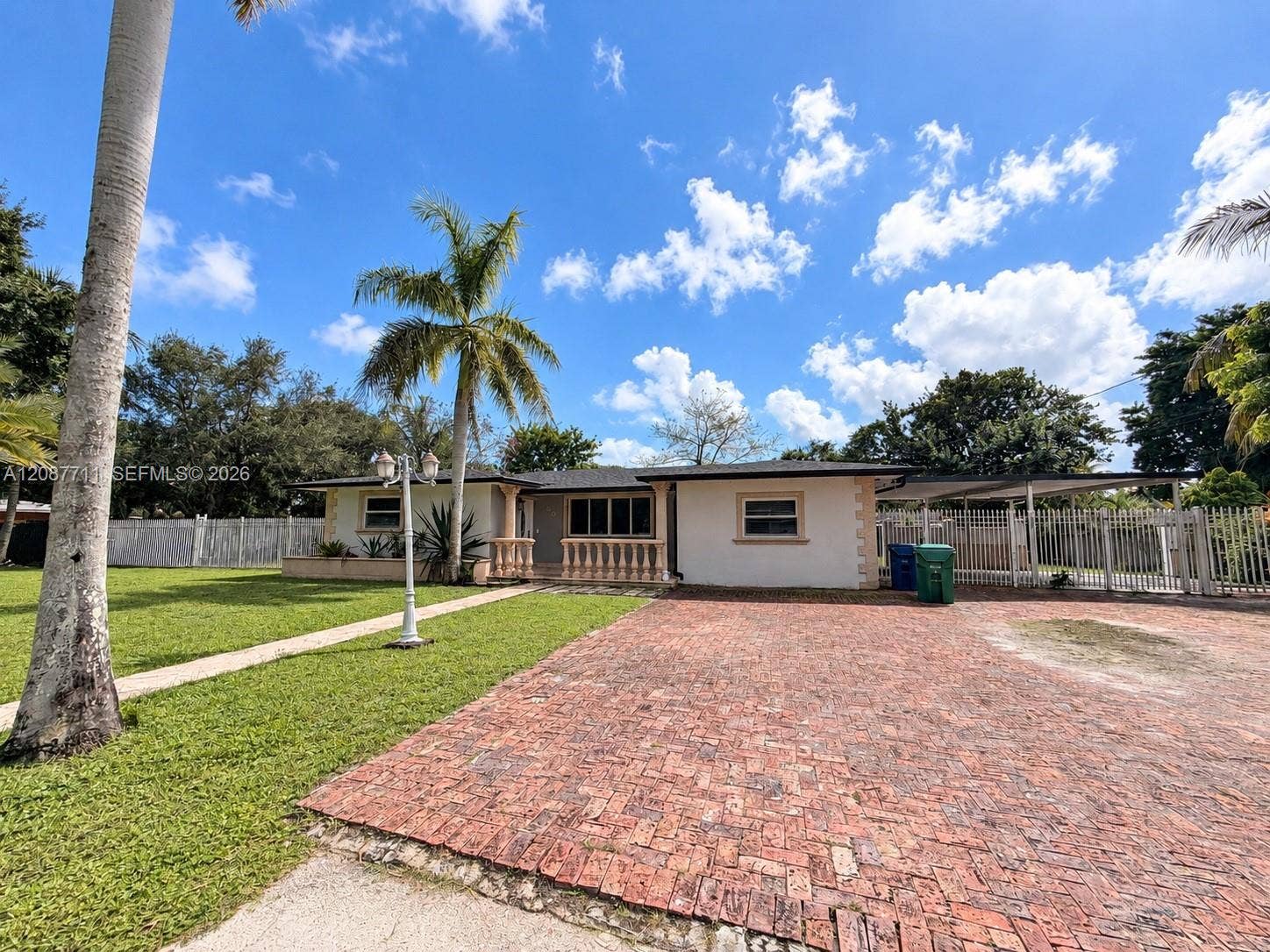 80 NE 162nd Street, Miami, Miami, FL 33162 Single-story exterior featuring a brick-paved driveway, decorative balustrade porch railing, attached carport, and perimeter metal fencing