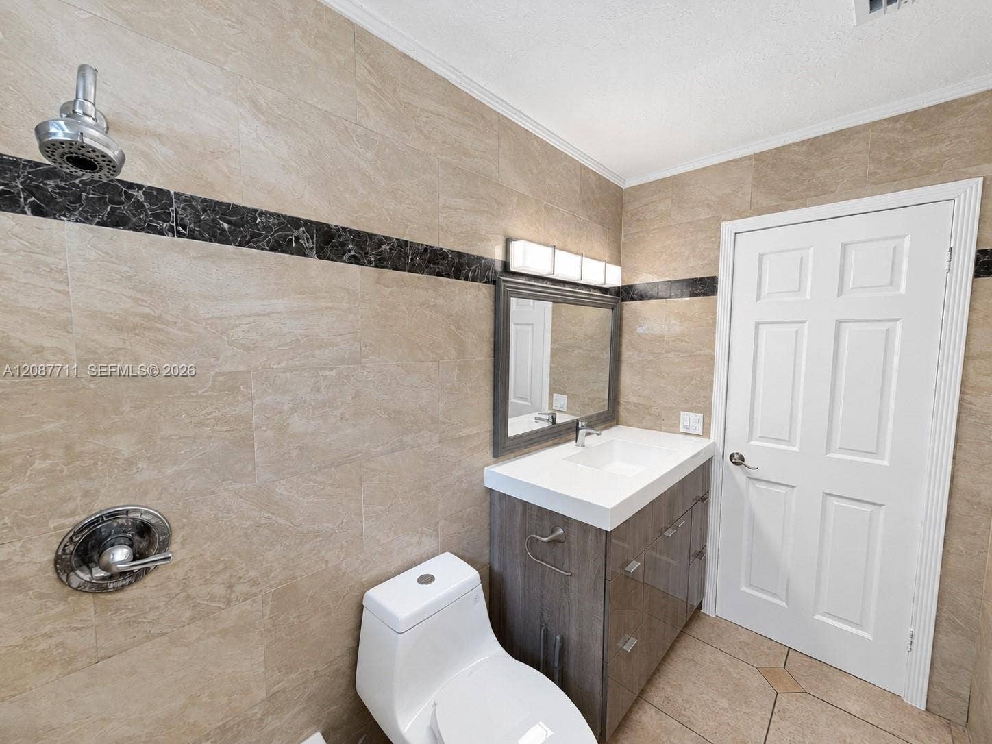 80 NE 162nd Street, Miami, Miami, FL 33162 Full-wall tile surround with dark stone accent banding, wood-finish vanity with integrated sink, four-light vanity fixture, and six-panel interior door