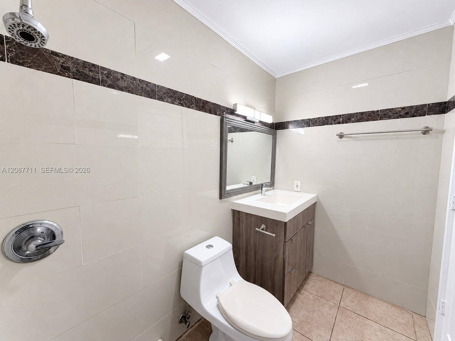 80 NE 162nd Street, Miami, Miami, FL 33162 Bathroom featuring light-toned wall tile with dark stone accent trim, wood-finish vanity with a rectangular sink, wall-mounted light fixture, and metal towel bar