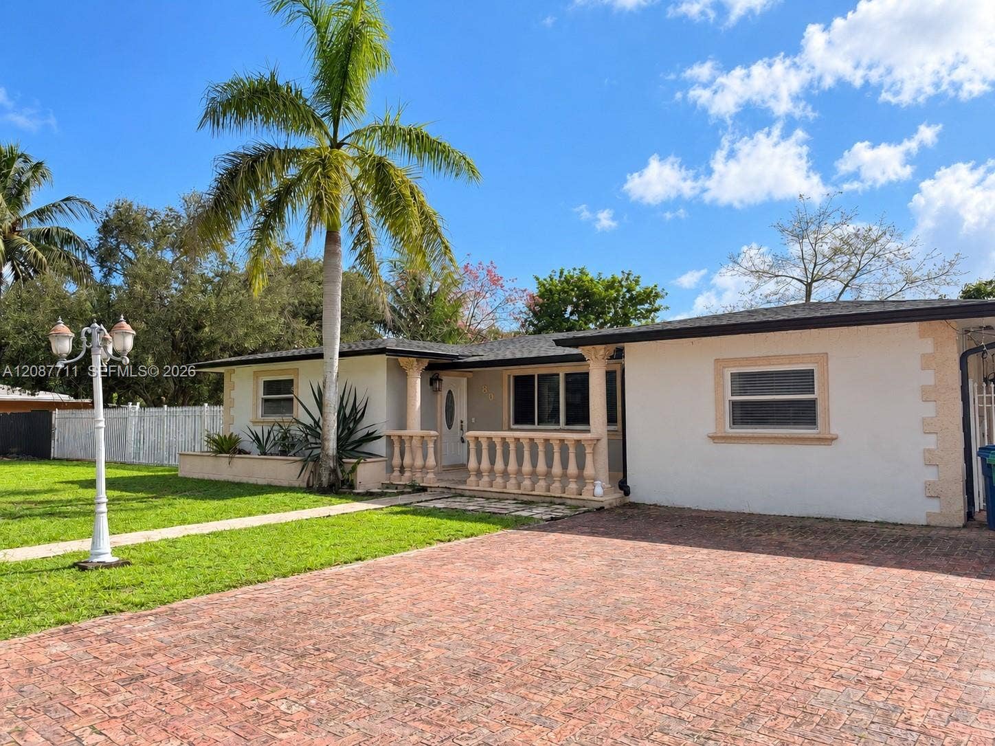 80 NE 162nd Street, Miami, Miami, FL 33162 Single-story exterior featuring a brick-paved driveway, decorative balustrade porch railing, stucco facade with stone-style corner quoins, and a pedestal post lamp