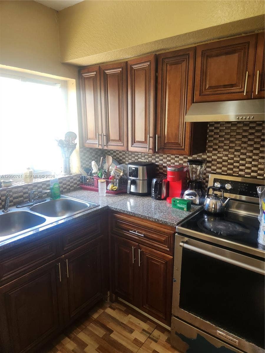 Kitchen featuring stained wood cabinetry, stone-finish countertops, stainless steel dual-basin sink, patterned tile backsplash, and stainless steel range