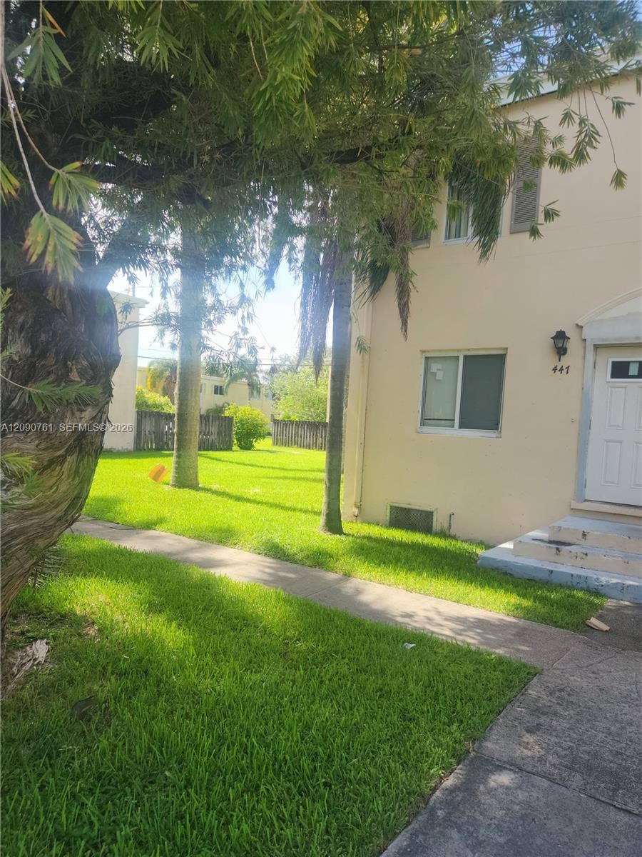 Exterior elevation featuring a stucco facade, concrete walkway, mature trees, and an enclosed grass yard
