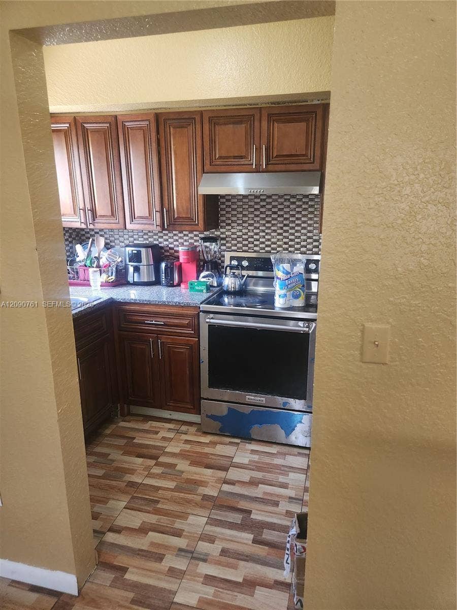 Kitchen featuring wood cabinetry, stone-finish countertops, stainless steel range with overhead vent hood, tiled backsplash, and patterned tile flooring