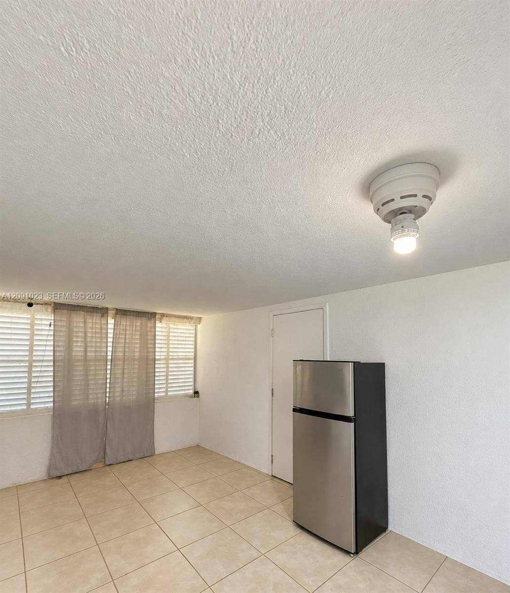Neutral-tone floor tiling, stainless steel refrigerator, white textured walls, and horizontal blinds