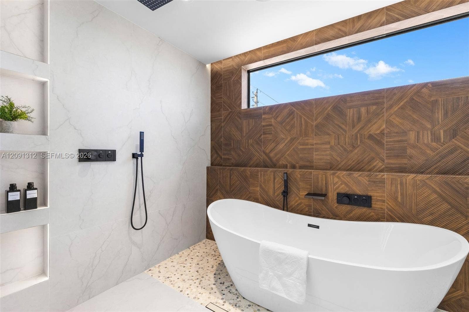 Freestanding soaking tub featuring matte black wall-mounted fixtures, geometric wood-patterned accent wall, recessed shelving, and a panoramic clerestory window