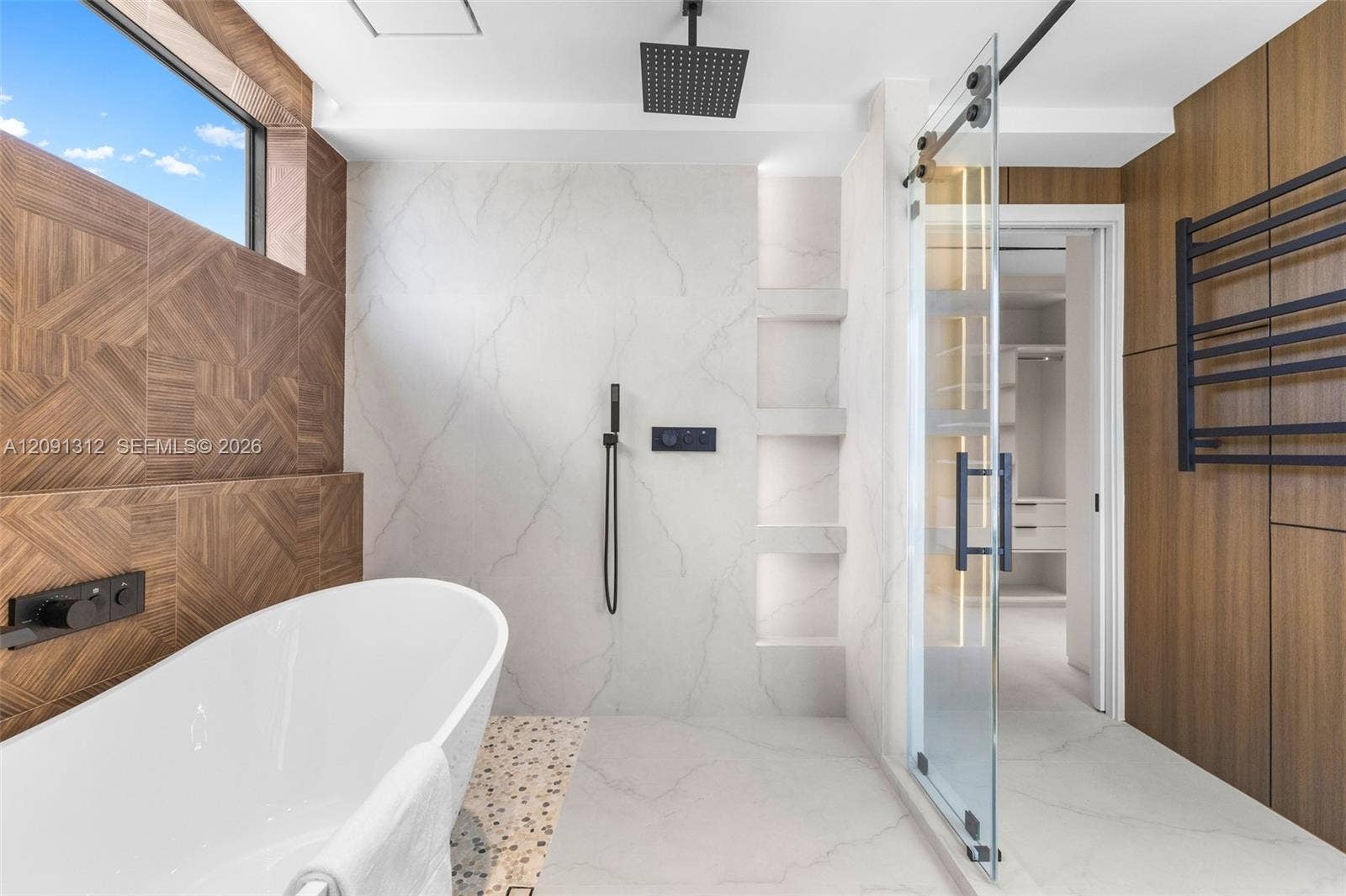 Freestanding soaking tub with matte black wall-mounted faucet, walk-in shower with glass sliding door, rainfall showerhead, integrated tiled shelving, and wood-finish wall accents