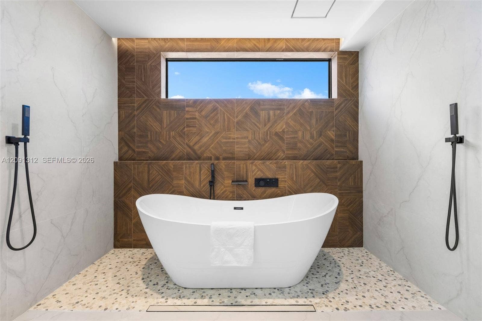 Freestanding soaking tub with matte black handheld shower fixtures, light-toned stone-finish tile surround, geometric wood-patterned accent wall, and horizontal clerestory window