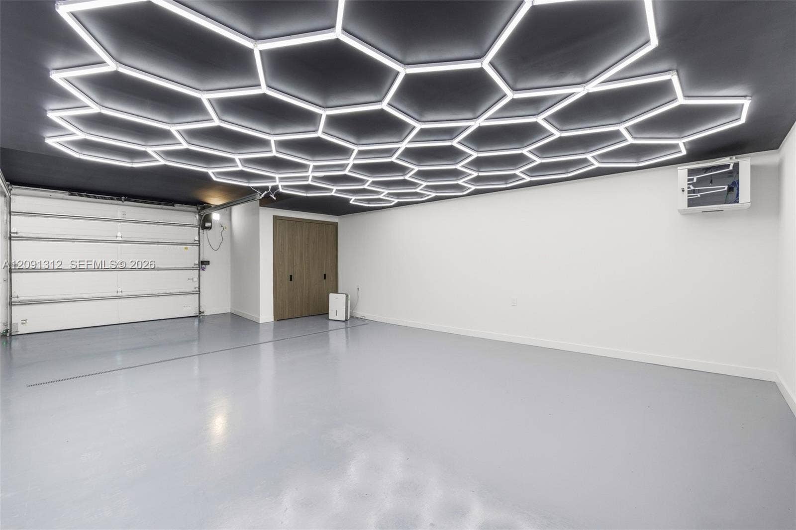 Custom ceiling with hexagonal LED light panels, smooth-finish gray flooring, white overhead garage door, and flat-panel wood-finish storage cabinetry