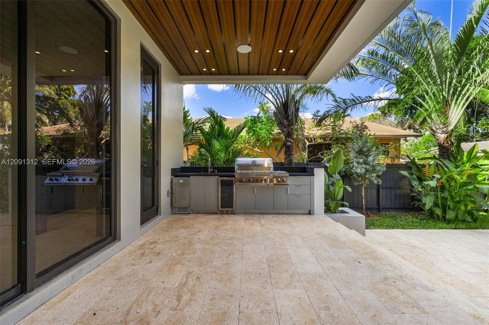 Covered outdoor kitchen featuring built-in cabinetry, stainless steel grill, beverage cooler, stone-finish flooring, and a wood-slat ceiling