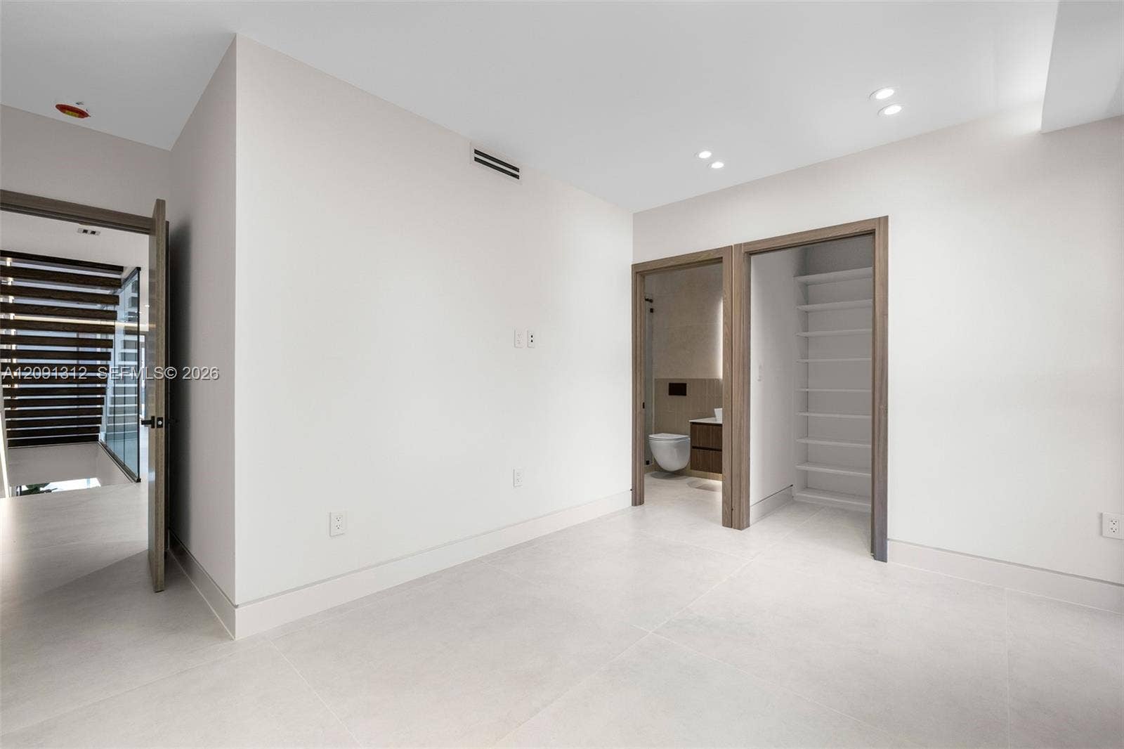 Neutral interior space featuring large-format tile flooring, recessed ceiling lighting, and wood-trim doorways leading to a built-in shelving unit and a bathroom