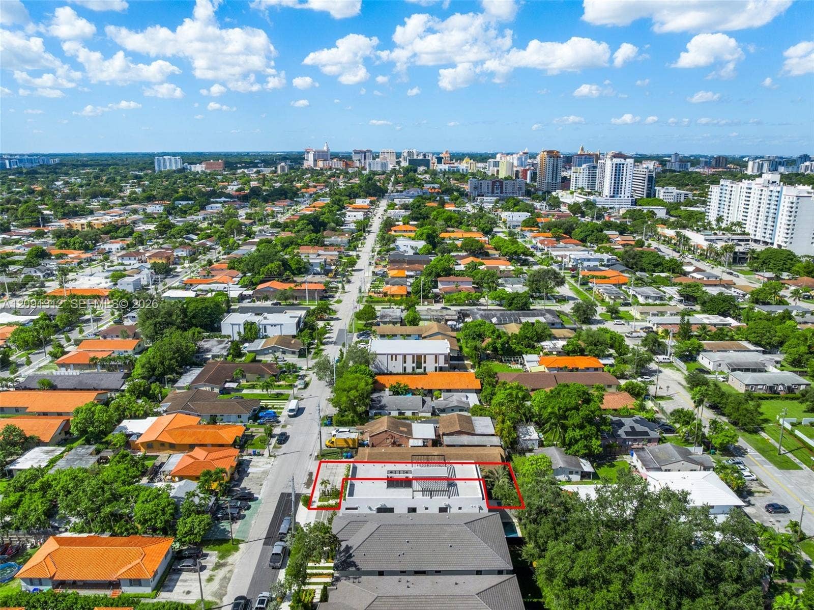 Aerial perspective of a multi-story property featuring a flat roofline, integrated rooftop terrace, ground-level paved surfacing, and mature perimeter landscaping within a dense urban landscape