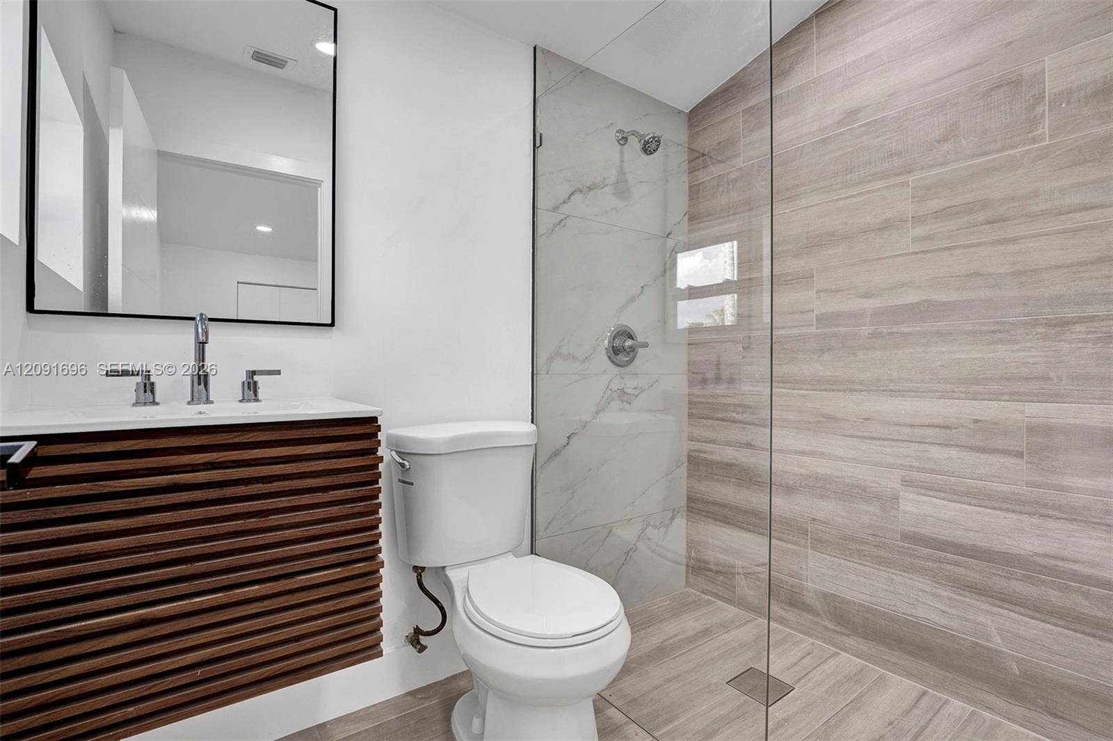 Modern bathroom featuring a wall-mounted slatted wood-finish vanity, large-format tile shower surround with a frameless glass enclosure, and a rectangular mirror with a black frame