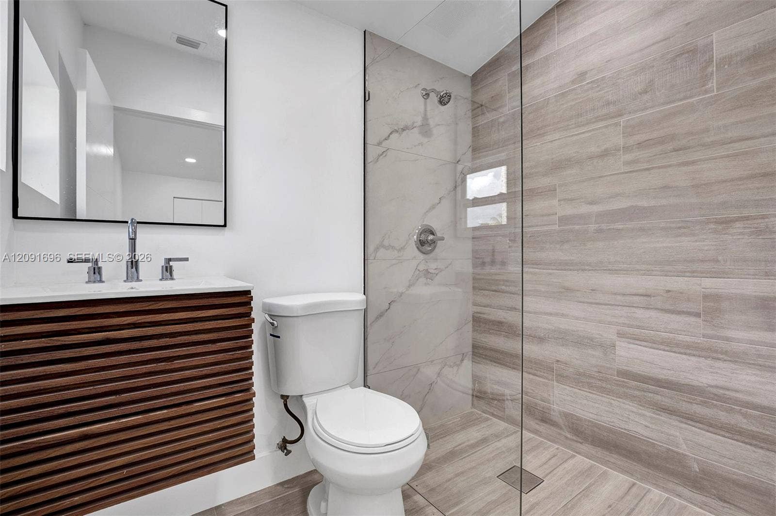 Bathroom featuring a slatted wood-finish floating vanity, rectangular sink with widespread faucet, wall-mounted mirror with a black frame, walk-in shower with a glass panel, and large-format tile surround