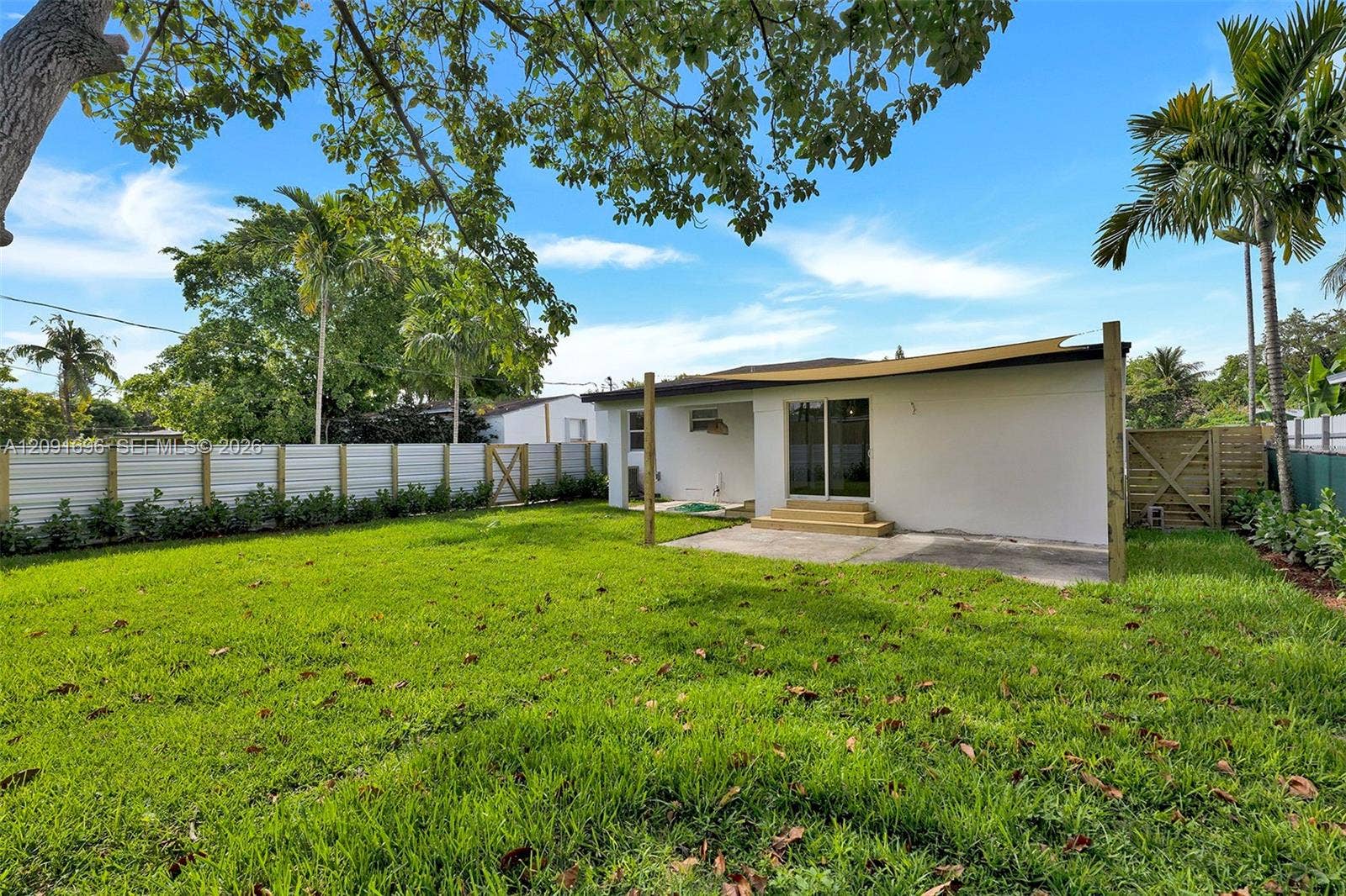 Fenced backyard featuring a manicured lawn, concrete patio, overhead sun shade structure, and mature palm trees