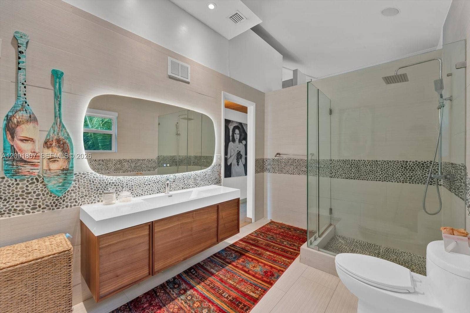 Floating wood-finish vanity with a white countertop and integrated sink, illuminated irregular-shaped vanity mirror, glass-enclosed shower with pebble tile accents, and large-format neutral wall and floor tiling