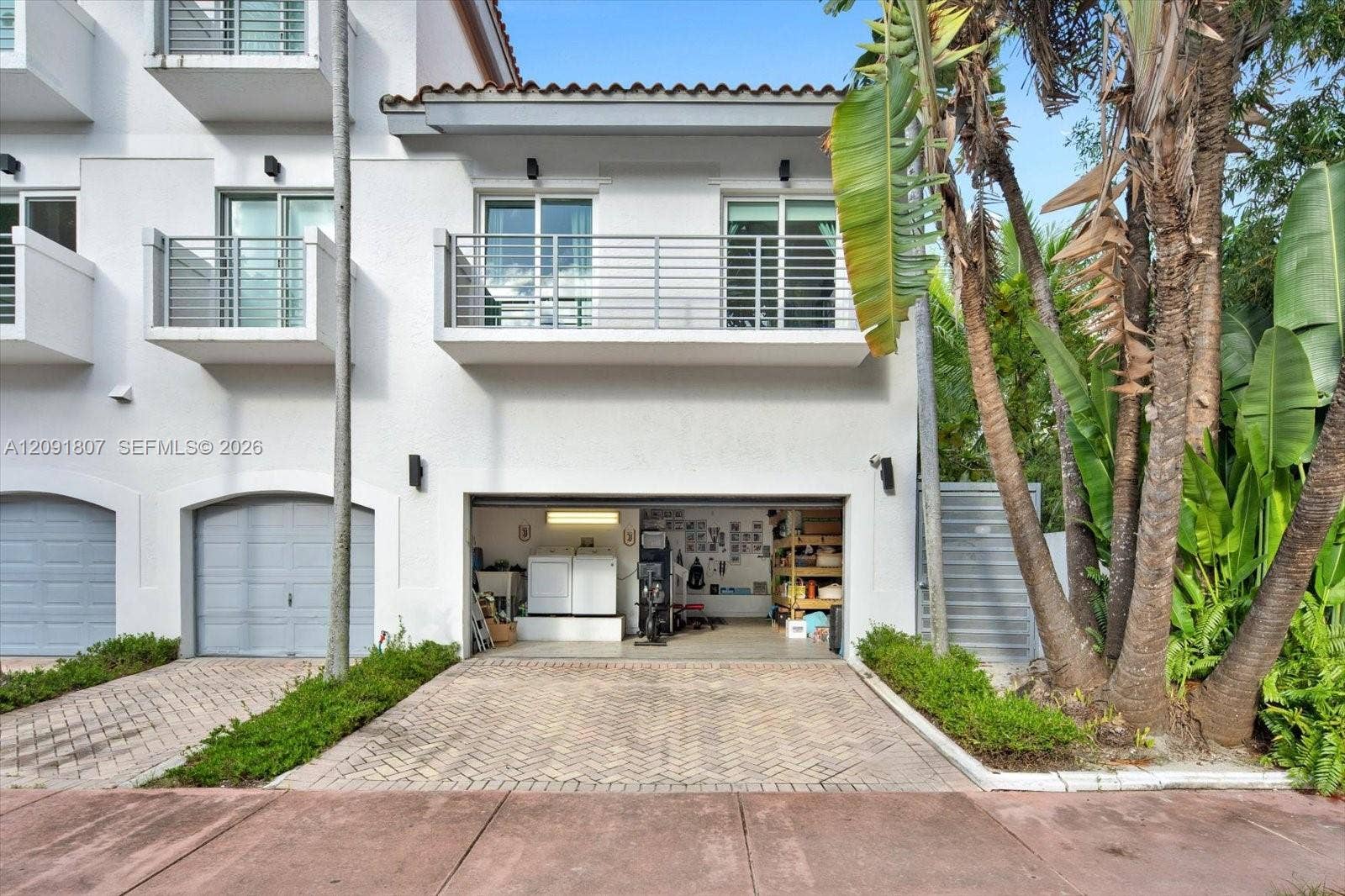 Stucco exterior featuring dual-level metal balconies, terracotta-style roof tile accents, a private herringbone-patterned paver driveway, and an attached multi-car garage