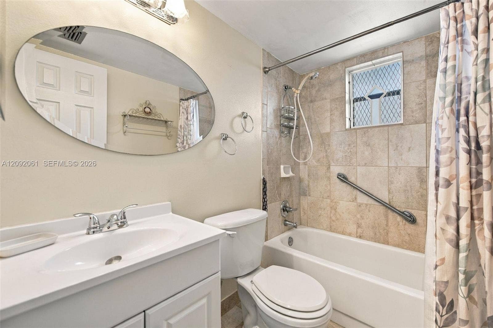 6555 W Broward Boulevard, #309, Plantation, FL 33317 Bathroom featuring a vanity with an integrated basin, an oval wall-mounted mirror, a tiled shower surround, a built-in grab bar, and a window with a lattice security grate