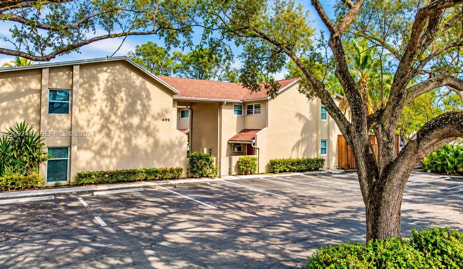 499 SE 13th Street, #7E, Fort Lauderdale, FL 33316 Some landscaping visually staged for illustrative purposes. Exterior building facade featuring a tan stucco finish, multi-pitched shingled roof, asphalt-paved parking area.