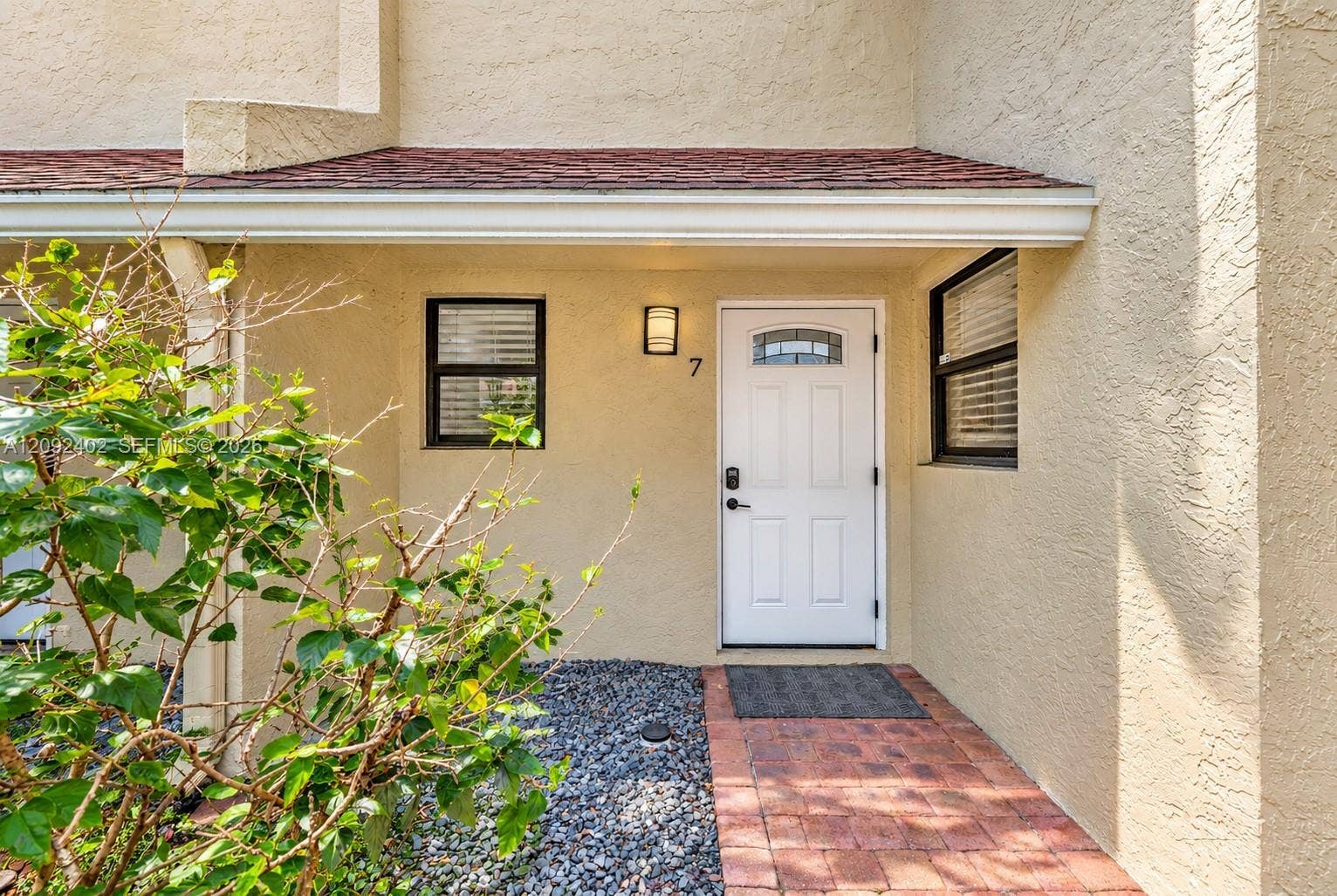 499 SE 13th Street, #7E, Fort Lauderdale, FL 33316 Stucco exterior entrance featuring a brick-paver walkway, red shingle roof overhang, white door with a glass inlay, and wall-mounted exterior lighting