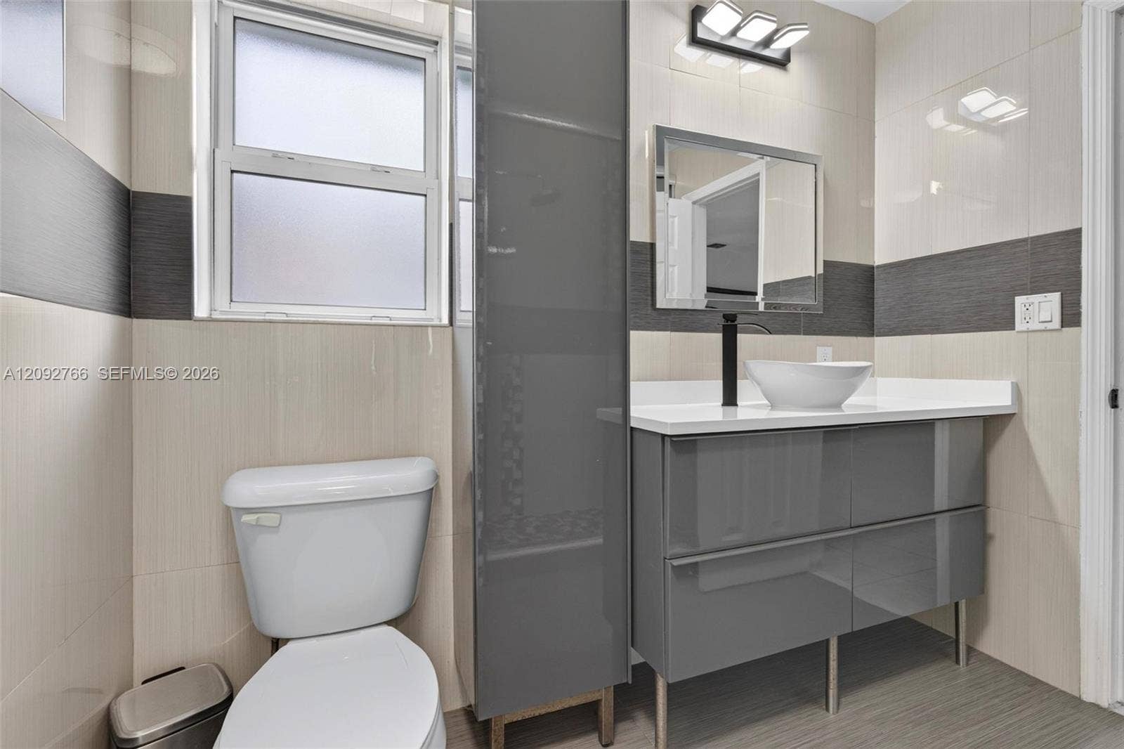Modern floating vanity featuring a glossy grey finish, white countertop, vessel sink, and matte black faucet, complemented by full-height wall tiling with contrasting horizontal banding