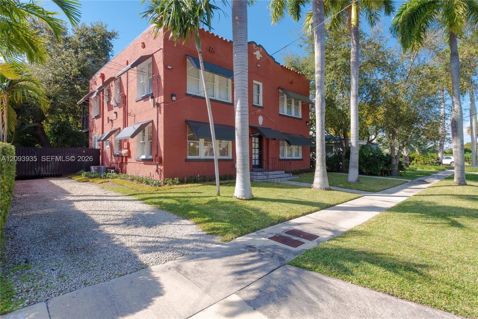 Two-story masonry exterior featuring mission-style architectural details, multiple metal window awnings, and a gravel parking area