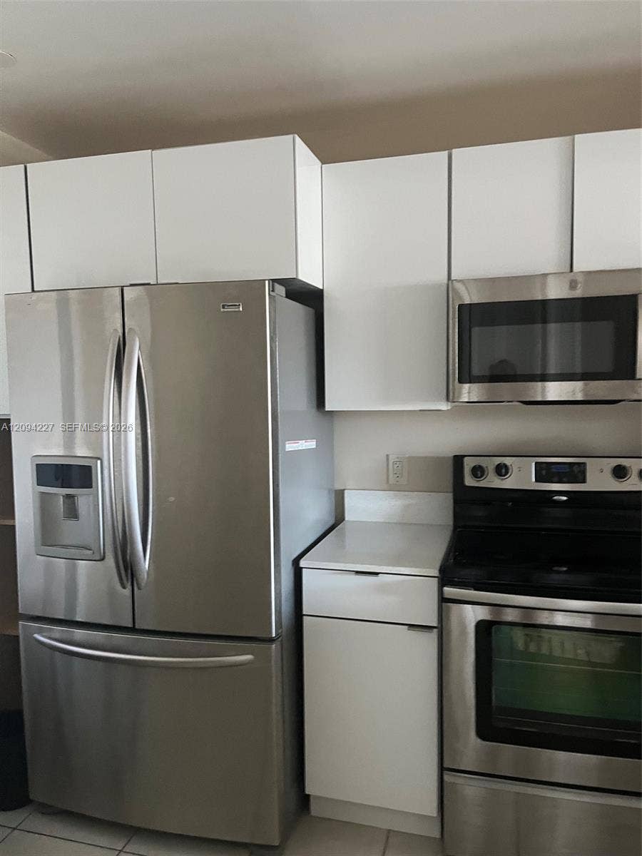 White flat-panel cabinetry featuring high-gloss finishes, stainless steel French-door refrigerator, matching stainless steel over-the-range microwave, stainless steel slide-in range, and white solid-surface countertops