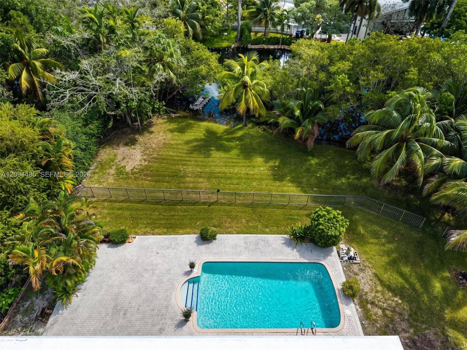 Outdoor living space featuring an in-ground swimming pool, concrete paver patio, metal perimeter fencing, manicured lawn, and mature tropical landscaping