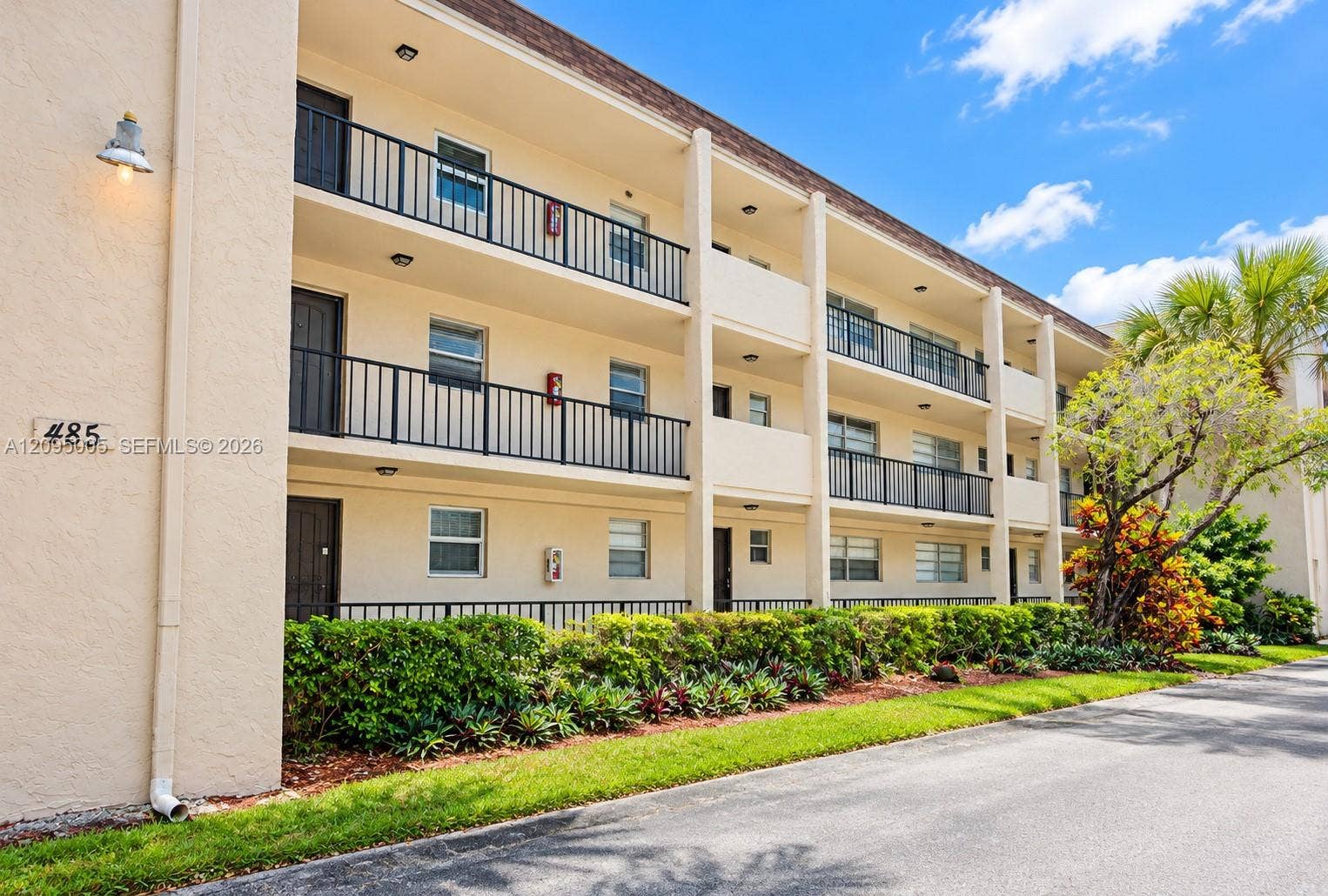 Three-story stucco exterior featuring stacked open-air balconies with metal railings, manicured foundation landscaping, and recessed exterior lighting