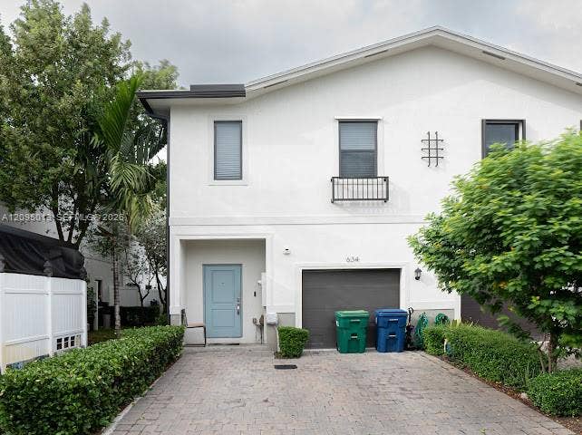 Two-story exterior featuring a light-toned stucco facade, a stone paver driveway, an attached garage, a window box, and a solid-panel front door