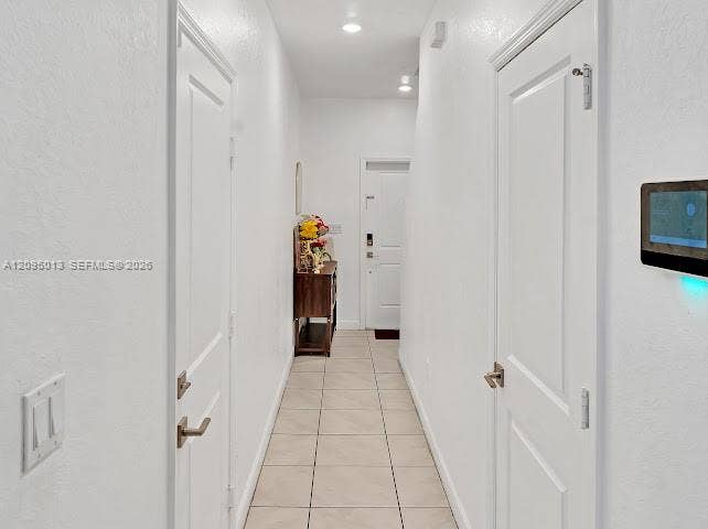 Hallway featuring neutral tile flooring, recessed ceiling lighting, wall-mounted smart home control panel, and solid-core interior doors with satin nickel lever handles