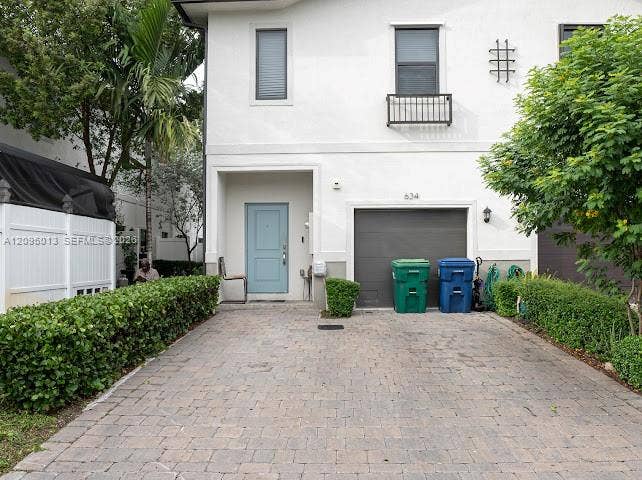 Stucco exterior featuring a paver driveway, an attached garage, and manicured privacy hedging