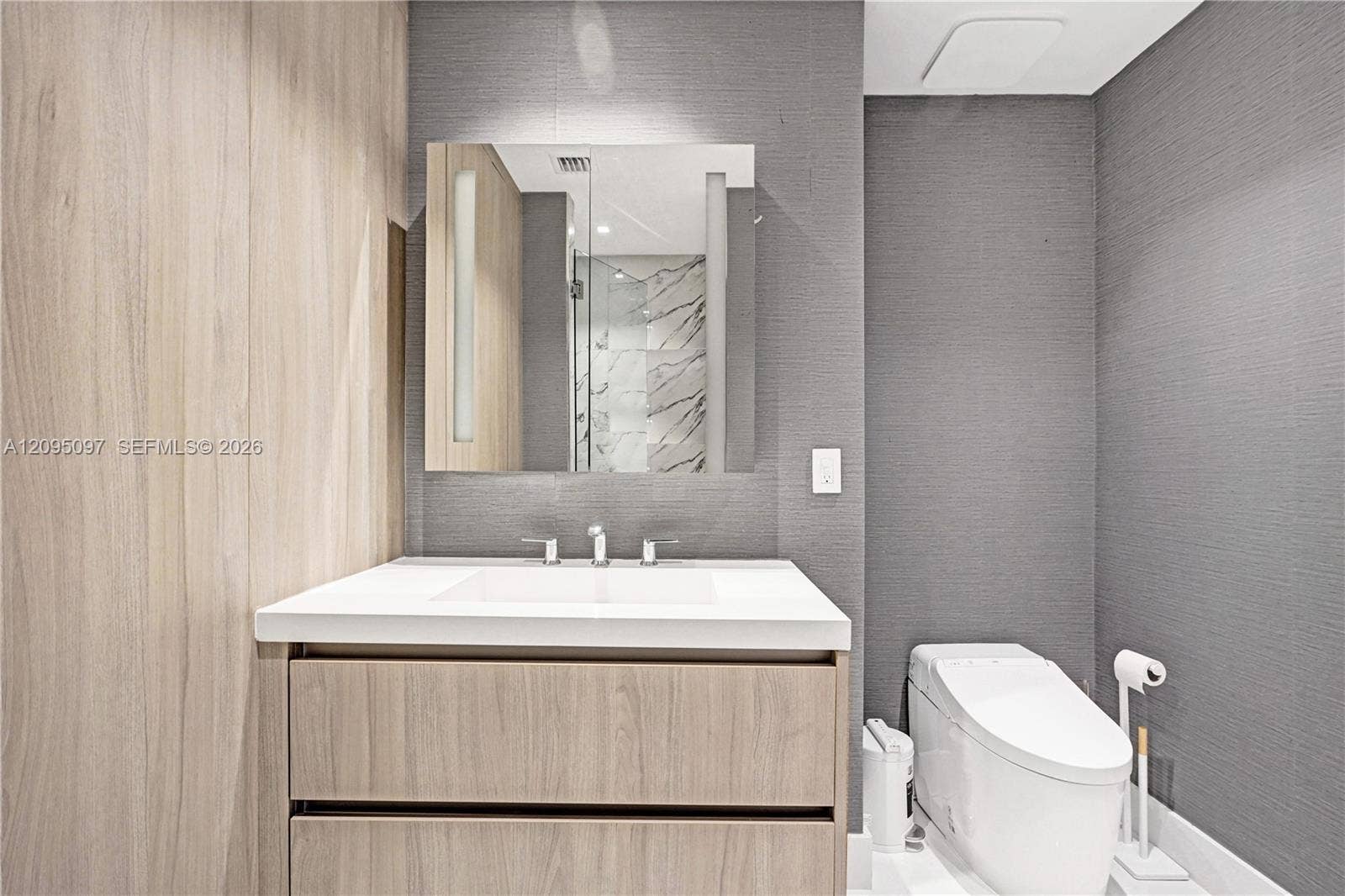 18201 Collins Avenue, #1407, Sunny Isles Beach, FL 33160 Floating vanity with wood-finish cabinetry, crisp white countertop, integrated rectangular sink, wall-mounted mirror with integrated lighting, and textured grey wall finishes