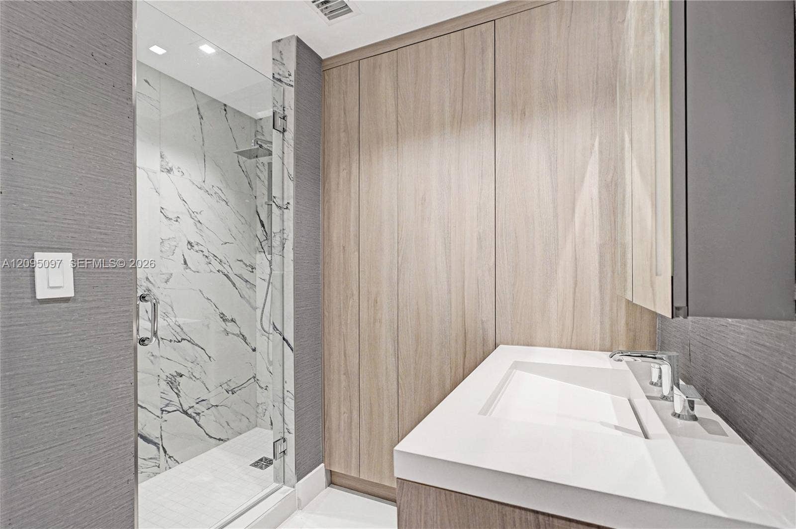 18201 Collins Avenue, #1407, Sunny Isles Beach, FL 33160 Modern bathroom featuring textured wall surfaces, integrated wood-finish cabinetry, a glass-enclosed shower with large-format veined tile, and a built-in vanity with a sleek solid-surface countertop and recessed basin