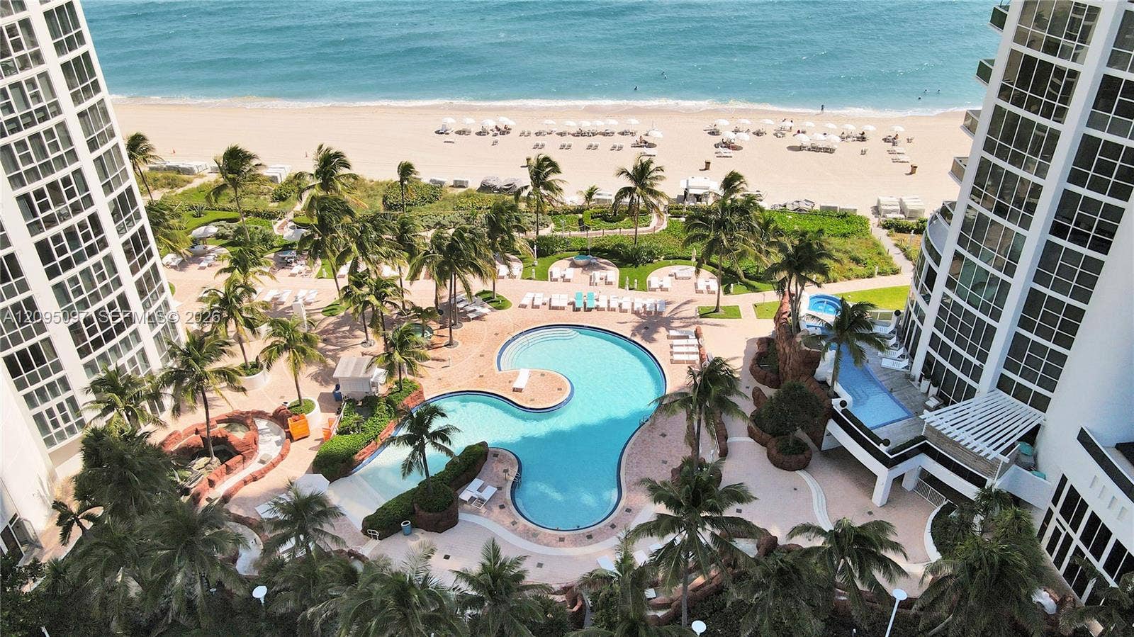 18201 Collins Avenue, #1407, Sunny Isles Beach, FL 33160 Aerial perspective of property grounds featuring a free-form swimming pool, integrated hot tub, paved sun deck, mature palm trees, and coastal access
