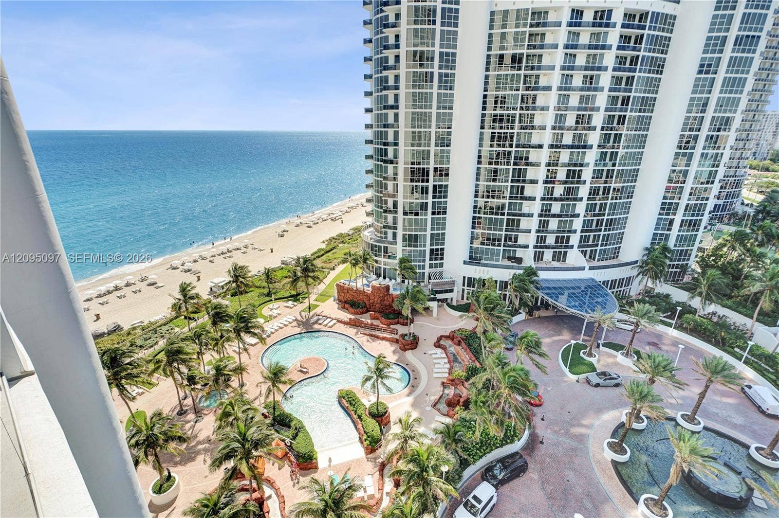 18201 Collins Avenue, #1407, Sunny Isles Beach, FL 33160 High-rise architectural exterior featuring glass-paned facades, a landscaped courtyard with a curvilinear swimming pool, mature palm trees, a circular driveway, and direct oceanfront positioning