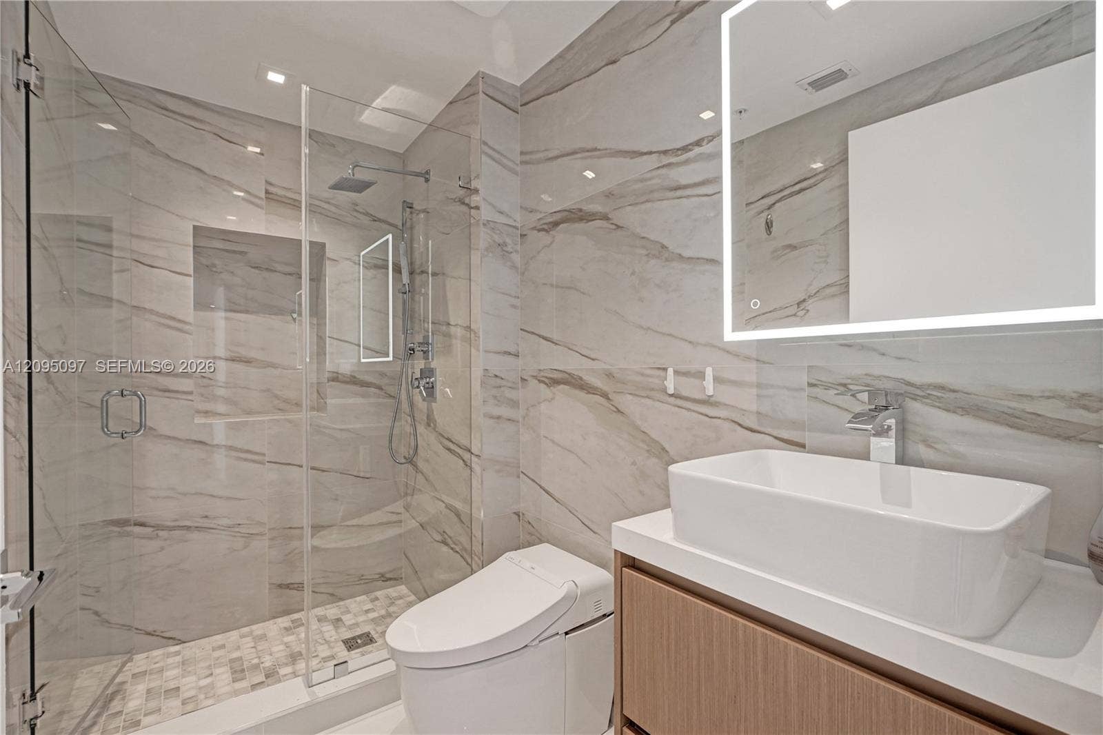 18201 Collins Avenue, #1407, Sunny Isles Beach, FL 33160 Bathroom featuring large-format wall tile, glass-enclosed shower with rainfall showerhead, built-in shower niche, floating wood-finish vanity with vessel sink, and illuminated wall-mounted mirror